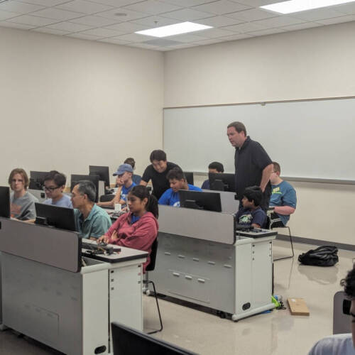Rows of people seated at computers in a classroom or computer lab, with a few instructors standing and assisting students. The setting conveys a learning environment focused on hands-on work and tech support